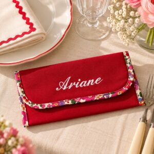 Pochette à serviette de table rouge avec biais Liberty fleuri et prénom Ariane brodé en blanc, présentée sur une table dressée.